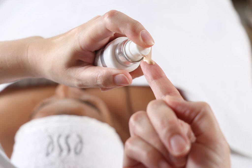 Close-up of hands applying a small amount of skincare product from a pump bottle onto a fingertip. In the background, a person with their eyes covered by a towel lies down on a treatment table, possibly in a spa or clinic setting.