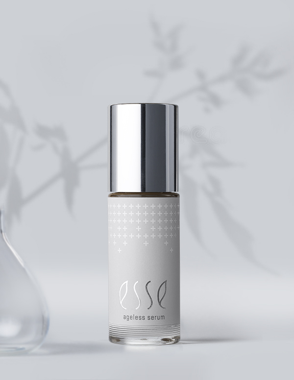 A bottle of Esse Ageless Serum with a shiny silver cap stands against a soft gray background, featuring blurred botanical shadows and part of a glass vessel on the left.