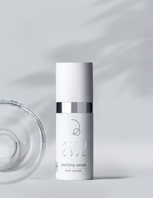 A white bottle labeled with "Esse Clarifying Serum" stands upright on a light gray surface. The bottle exhibits a sleek, minimalist design, accented by a silver band and distinctive black and green text. A shadow and part of a glass bottle are visible on the left.