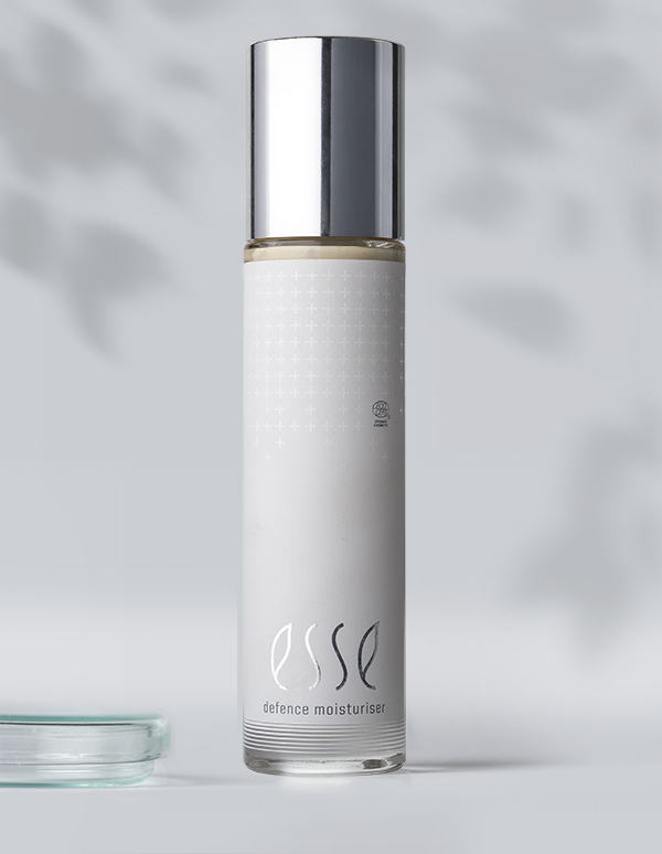 A sleek white bottle with a silver cap labeled "Esse Defence Moisturiser" stands upright against a soft, blurred grey background. Part of a clear glass dish appears in the lower left corner.