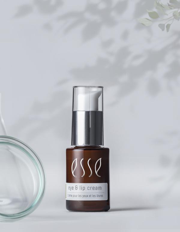A brown bottle with a silver pump dispenser labeled "Esse Eye & Lip Cream" is set against a soft, blurred background adorned with delicate leaves in the upper right corner, accompanied by a glass jar lying on its side beside the bottle.