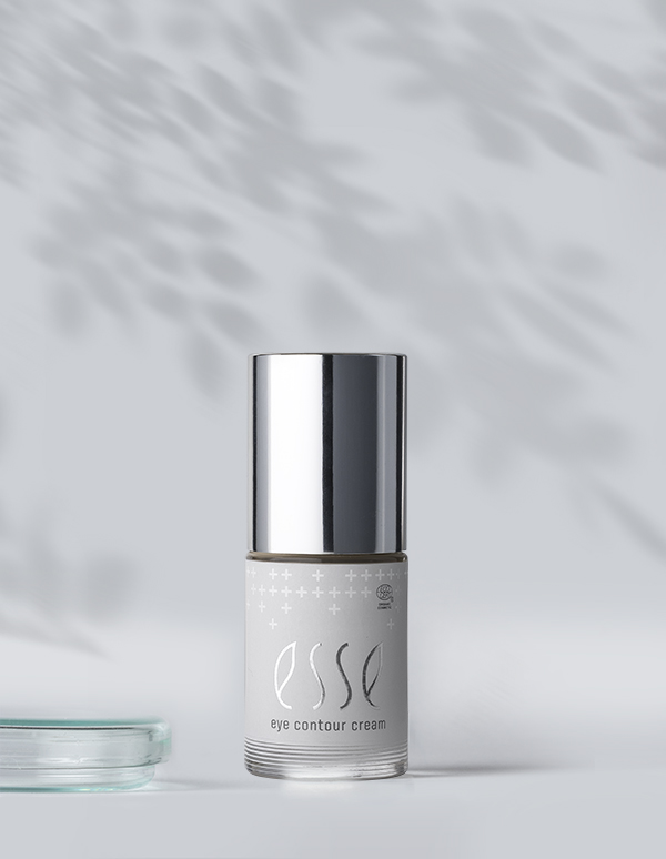 A small bottle of Esse Eye Contour Cream with a shiny silver cap rests on a light surface, soft leaf shadows in the background.