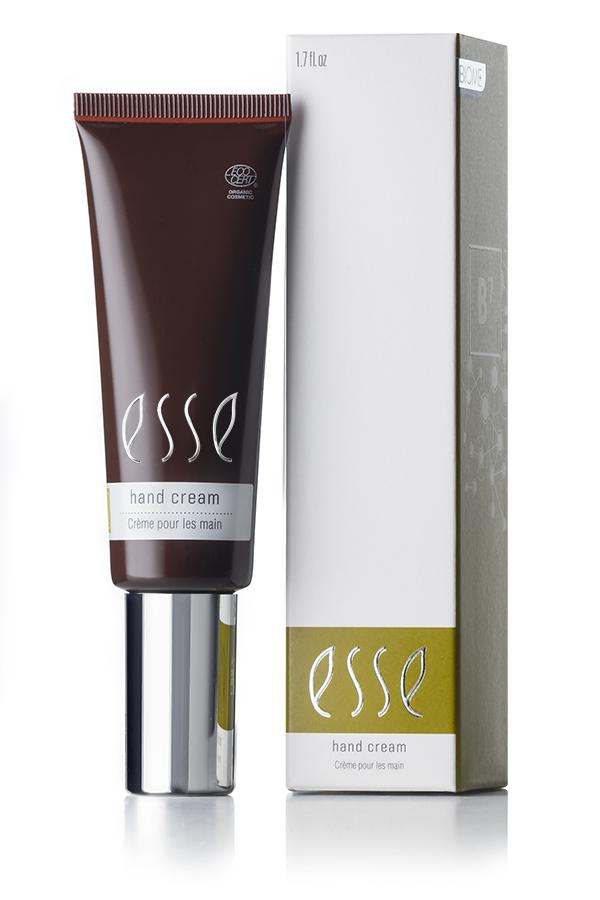 An Esse Hand Cream tube is positioned next to its corresponding box. The cream's tube is brown with a silver cap, featuring the "Esse Hand Cream" text in white. The box shares similar branding and colors, with additional product information.