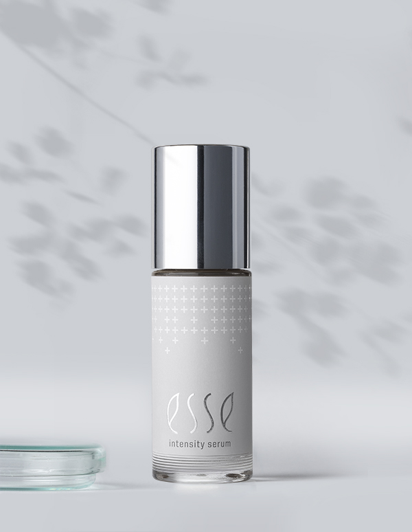 A bottle of Esse Intensity Serum with a shiny silver cap stands upright on a light surface, subtle leafy shadows falling across the pale gray background.