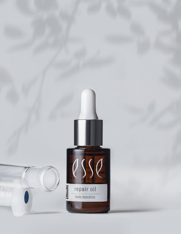 The "Esse Repair Oil" brown glass bottle with a dropper cap is positioned on a white surface. Leaf shadows cast in the background, alongside part of a transparent cylindrical item partially visible near the bottle.