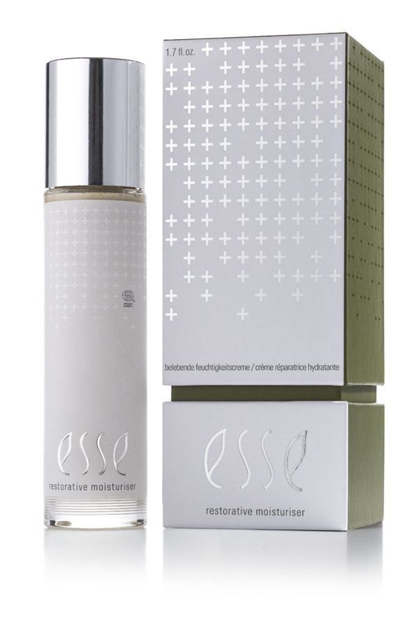 A bottle of Esse Restorative Moisturiser with a metallic cap stands beside its matching silver and green box, both showcasing a minimalist design with white cross patterns and the Esse logo.
