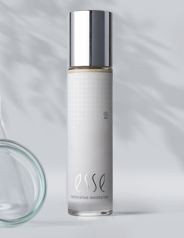 A sleek white bottle with a silver cap labeled "Esse Restorative Moisturiser" stands upright on a light gray surface, soft shadows cast around it and a subtle glass object nearby.