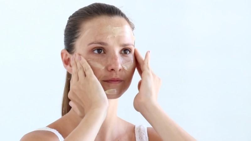 A woman is diligently applying the Award-Winning Esse Cream Mask to her cheeks and forehead with both hands. With a focused expression, she is dressed in a white tank top, set against a plain, light-colored background.
