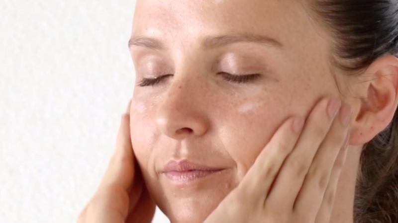 A person with closed eyes gently touches their face with both hands, showcasing a serene expression. The softly lit background enhances the smooth and relaxed appearance of their skin, thanks to the Esse Defence Moisturiser.