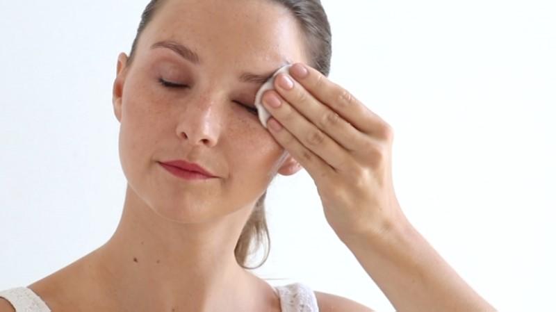 A person with an updo hairstyle is cleansing their face using a cotton pad soaked in Esse Make-up Remover. They have their eyes closed and appear to be gently wiping their forehead. The background is plain and white, highlighting the person's relaxed expression and skincare activity.