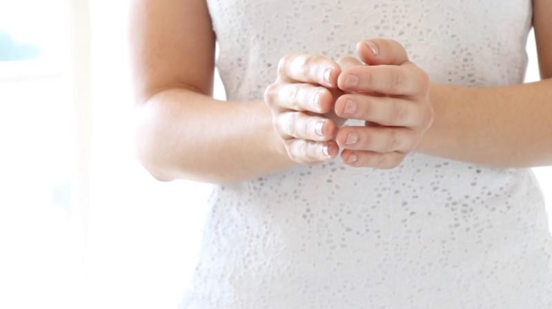 A person is using Esse Protect Oil while wearing a white lace dress and standing with their hands clasped together at waist level. The background is softly lit and out of focus.