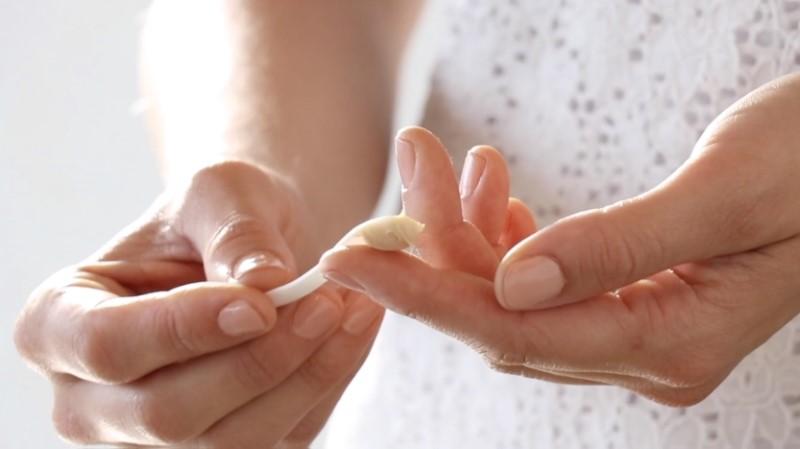 A person in a white lace shirt applies Esse Rich Moisturiser from a white tube onto their fingertip. The image focuses on the hands and forearms, capturing the action of dispensing and preparing to use the moisturiser. The background is softly blurred.