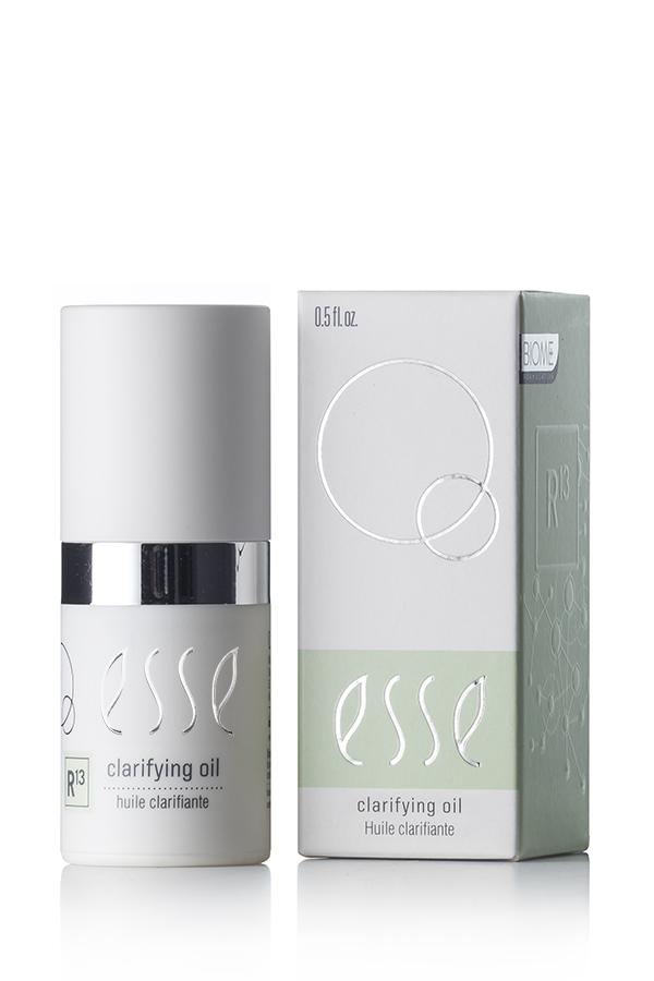 The image features a bottle of Esse Clarifying Oil along with its packaging. The cylindrical bottle is adorned with a white cap and a metallic silver band. The light green and white box showcases the product name and various branding elements. The bottle has a capacity of 0.5 fl oz.
