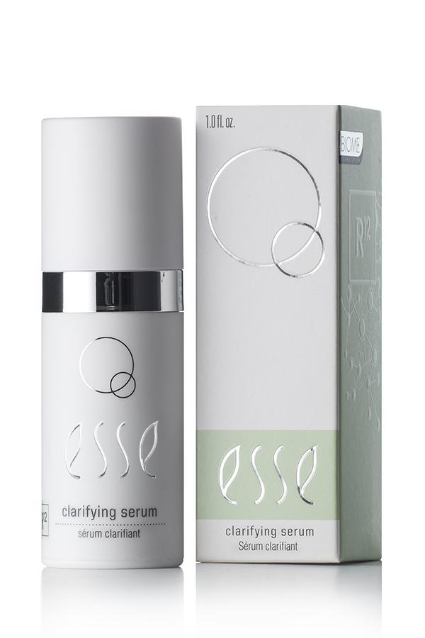 A 1.01 fl. oz. bottle of Esse Clarifying Serum is displayed alongside its packaging. The bottle, which is white with a silver cap, bears a label adorned with elegant circular designs. The packaging mirrors the design of the bottle and includes additional informational text.