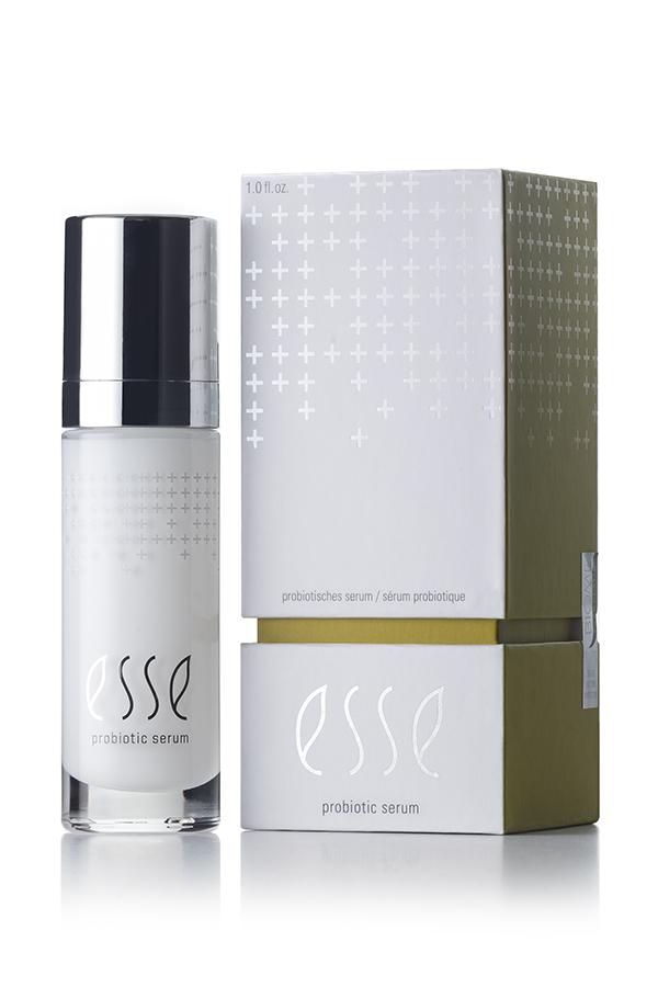 An Esse Probiotic Serum sits beside its corresponding box. The bottle is white with silver accents and a black cap, proudly showcasing the "esse" logo. The similarly white box sports a minimalist design adorned with small cross symbols and the "esse" logo.