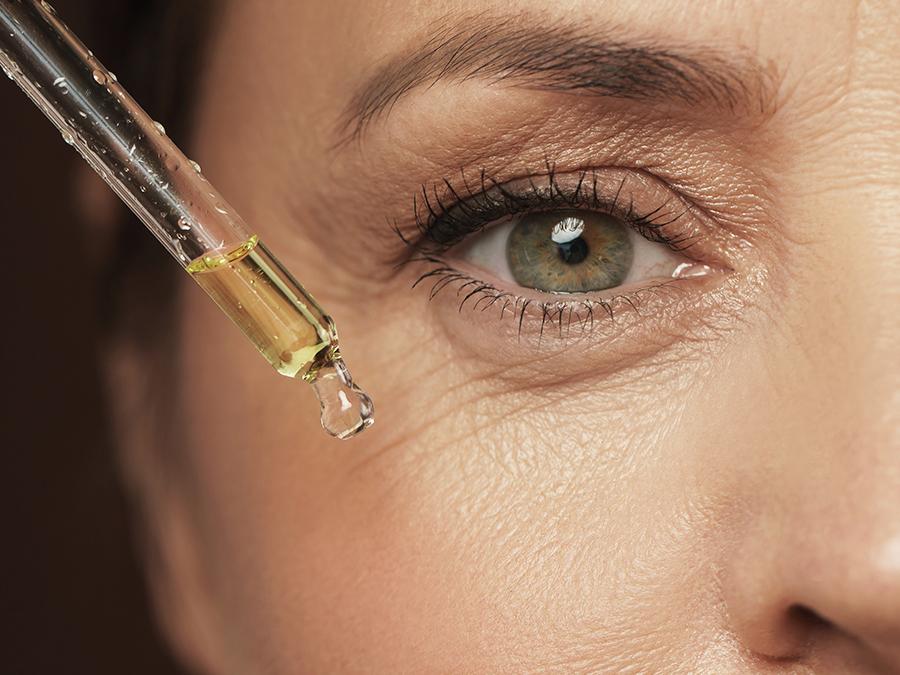 Close-up of a person's eye and part of their face. The individual is holding a dropper releasing a drop of yellow liquid onto the skin near the eye. The skin around the eye shows fine lines and wrinkles. The background is blurred, focusing attention on the eye and dropper.