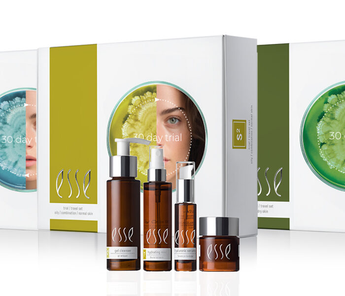 A set of Esse skincare products, including bottles and a jar, is displayed in front of three Esse 30 Day Trial Travel Sets, each featuring botanical designs and partial faces on the packaging.