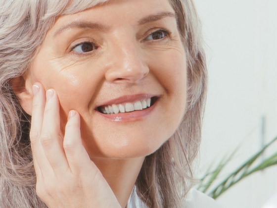 A smiling older woman with light skin and gray hair touches her cheek with her hand, radiating joy. A green plant is visible in the blurred background, evoking the warmth of a Conscious Gifting Festive Set.