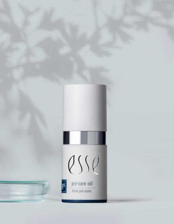 A white bottle of Import placeholder for 51695 pre-care oil with a silver band sits on a light surface. Leaf shadows fall across the background, adding a calming, natural feel. A clear glass dish is partially visible to the left.